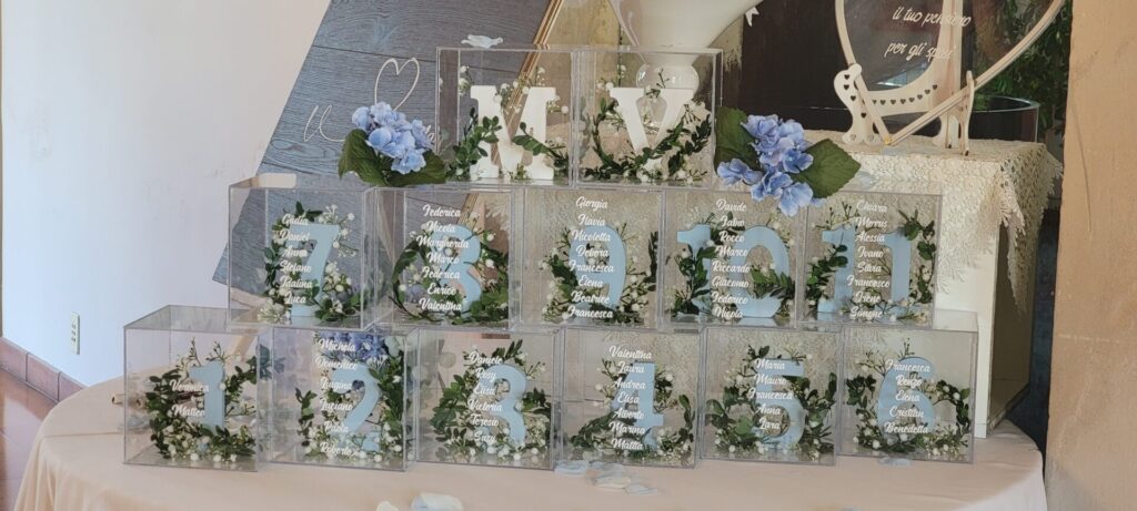 Modern acrylic wedding seating chart with white calligraphy, lush greenery, and soft blue hydrangea accents.